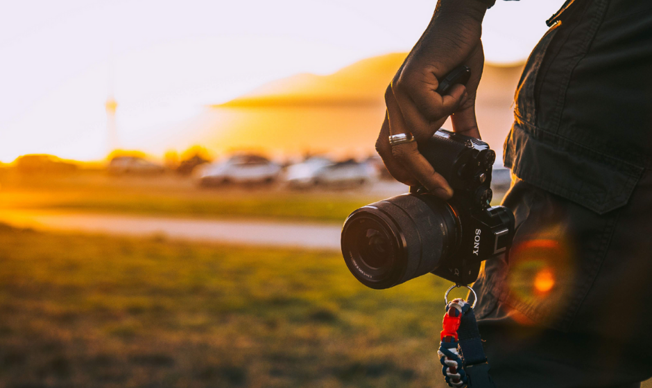 Close up of a back view of a hand holding a camera. In the background is a view of a park area with cars parked that are blurred out.