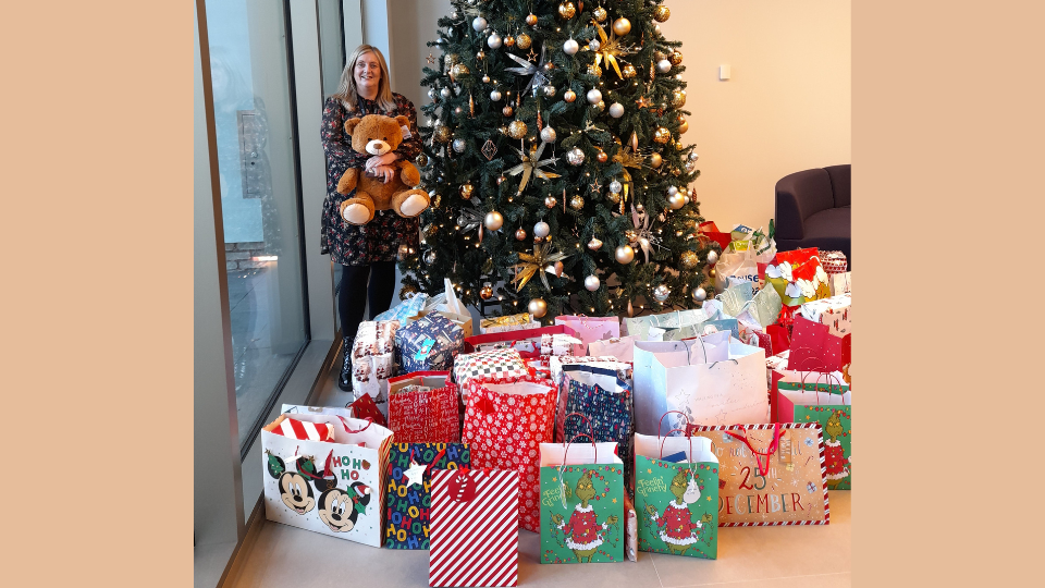 Kim, from BT/EE group poses with a large teddy bear, by a christmas tree and surrounded by presents donated by BT/EE staff for young carers in Dundee.