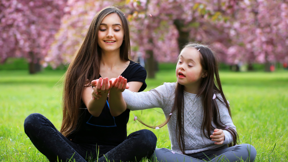 Image of a young girl and a little girls sitting cross legged on grass. The young woman has an object in her hand and the younger girl is looking at it.
