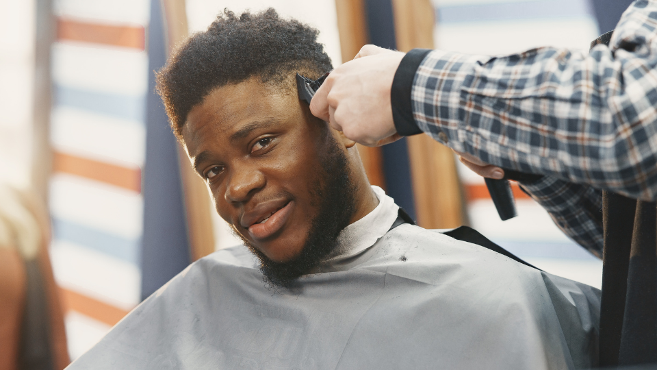Young man sits in a barber and has his head tilted, looking at the camera while getting his hair shaved at the sides by a barber that is off camera.