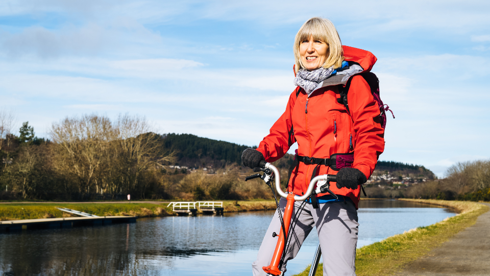 Woman wearing a red weatherproof jackets sits on a bike. In the background is a view of a small loch/lake.