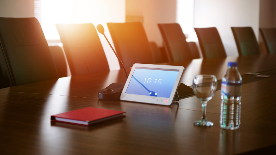 Image of a boardroom table with sun steaming through winder. A tablet with the time 10.15 propped up on the table along with a conference mic, bottle of water and a wine glass.