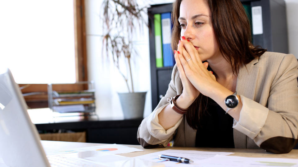 Woman sitting at a desk, looking pensive with fingers steepled. She appears to be looking at a laptop.