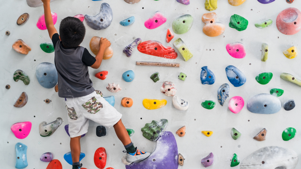 Close up view of a bouldering/climbing wall with lots of different coloured footholds. To the left is a young boy who is climbing the wall and is reaching up to a foothold off camera.