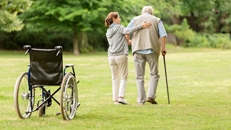Backview of an elderly man with a walking stick being helped by a young woman to walk across a garden/park. In the foreground there is an empty wheelchair.
