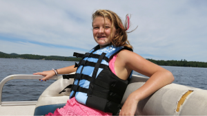 Teenage girls wearing a blue life jacket. She is sitting in a relaxed fashion in the back of a sailing boat. In the background is a view of a lake