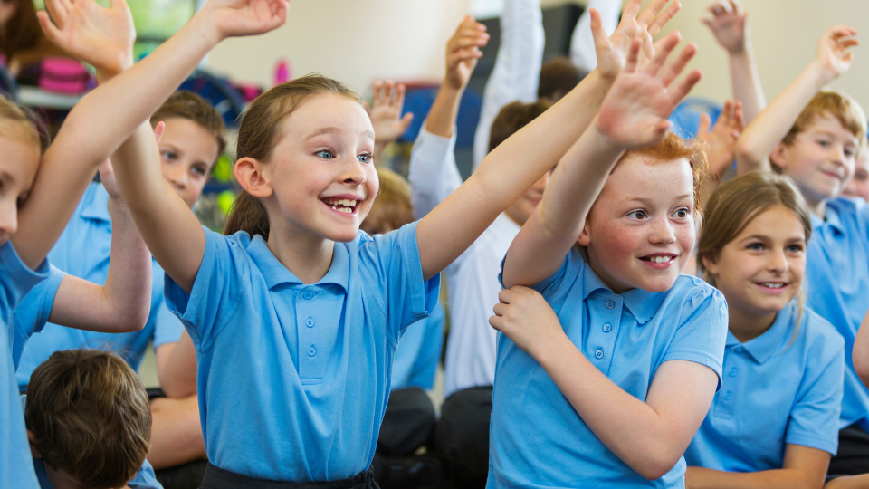 Close up view of a group of school children sitting looking off camera. All the children are wearing blue polo shirt and have their arms raised as if to answer a question.