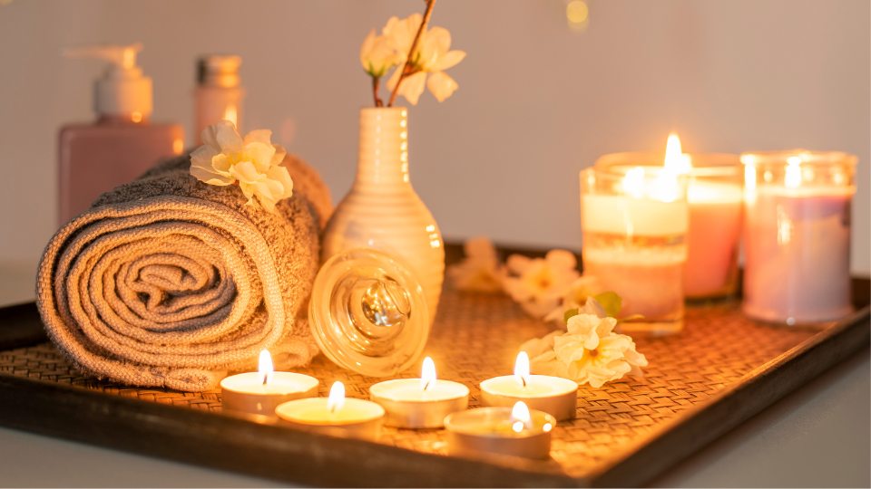 Close up of a tray. On the tray is a couple of beauty products, rolled-up towel, small vase with flowers, three lit jar candles and lit tealights in the foreground.