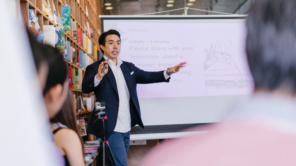 candid photo of a gentleman standing mid-presentation. His arms are spread wide to emphasise a point he is makking.