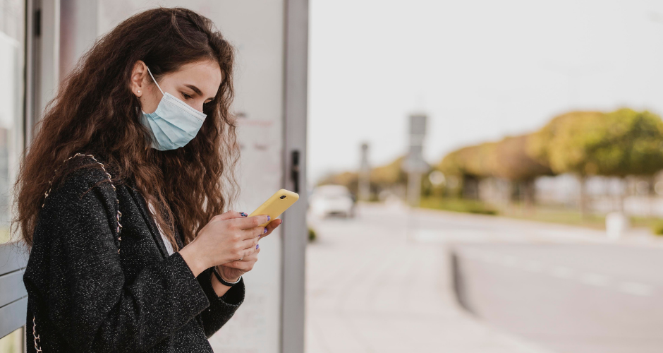 Side view of a young woman wearing a facemask looking at her mobile device at a bus stop