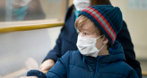 Young boy wearing a wool hat a facemask sitting on a bus looking out of the window