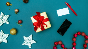 Blue background with Christmas decorations and a white preent with a red ribbon and a mobile phone strewn across.