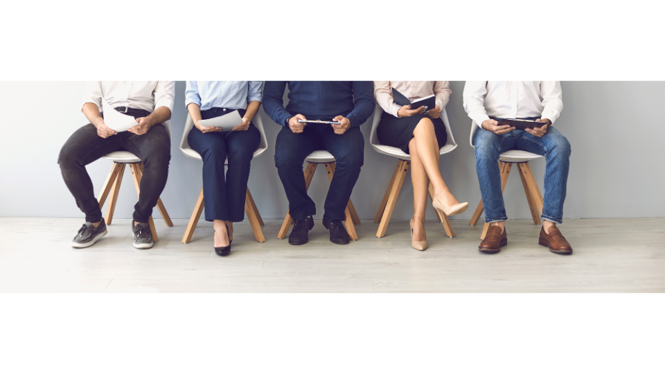 view of legs sitting in a row of chairs. Some have their hands in the lap and some are holding sheets of paper.