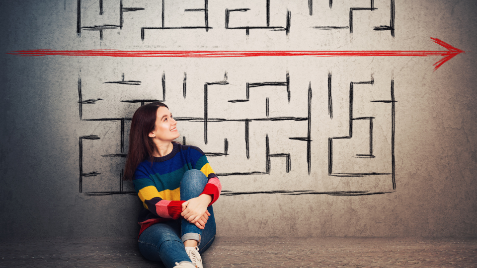 Young woman sitting cross-legged smiling up at a drawing of a maze drawing with a large red arrow drawn right through the middle.