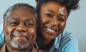 A photo of a man and a younger woman the woman appears to be embracing the man from behind. Both are smiling.