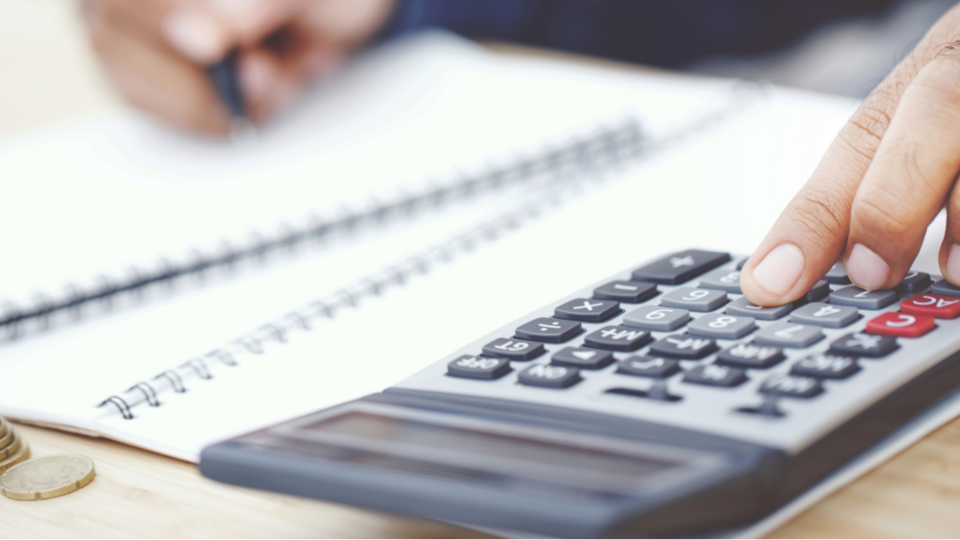 Close up view of a mans hands one had is typing on a calculator - the other is holding a pen and writing on a notebook.