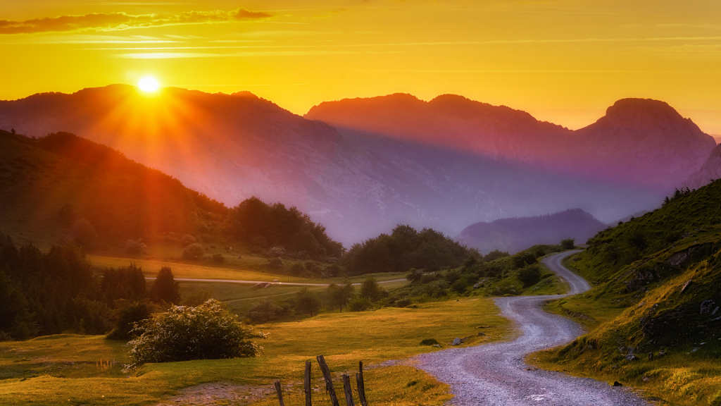 Image of a sunset against a range of mountains. In the foreground there is a curving road leading to the mountains.